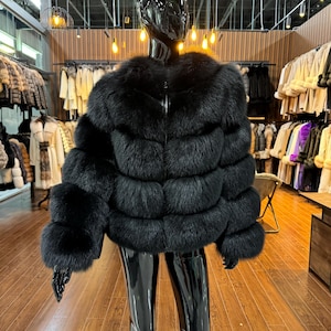 Puede incluir: Chaqueta de piel negra exhibida en un maniquí. La chaqueta presenta un cuello redondo y paneles horizontales de piel. El maniquí lleva pantalones y guantes negros. El fondo muestra una tienda de ropa con varios abrigos de piel.