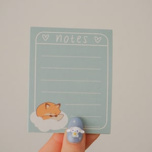 May include: A pale blue notepad with the word "notes" at the top, framed by hearts. A cartoon fox on a cloud is at the bottom. The notepad is held by a hand with a blue nail with a cartoon character.