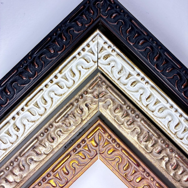 Op de afbeelding: Close-up van vier sierlijke hoeken van fotolijsten in zwart, wit, brons en goud. Elk frame heeft een gedetailleerd, herhalend scrollontwerp. De frames zijn gerangschikt in een gelaagde, schuine compositie tegen een witte achtergrond.