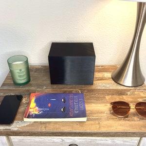 May include: A black box, a smartphone, a copy of "The Great Gatsby", sunglasses, and a green candle are on a wooden bedside table. A silver lamp with a white shade is also present. The table has a distressed wood finish.