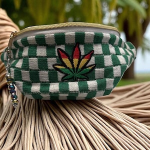 May include: Green and white checkered fanny pack with a gold zipper and a colorful cannabis leaf patch. The bag has a small beaded charm attached to the zipper. The fanny pack is made of a woven material.