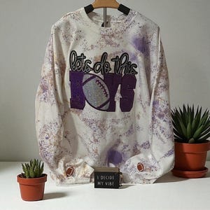 May include: White and purple tie-dye sweatshirt with the text "lets do this BOYS" in purple and silver. The sweatshirt is hanging on a wooden hanger. A small sign reads "I DECIDE MY VIBE".