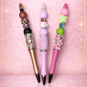 May include: Three decorative pens with beaded tops. One pen is gold with a black tip and a beaded top of brown, leopard print, and black beads. The second pen is light purple with a beaded top of pink and white floral beads. The third pen is pink with a beaded top of sprinkles.