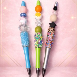 May include: Three decorative pens with colorful beaded designs. One pen has a blue barrel and a light blue, glittery grip. The second pen has a green barrel and a green, sprinkle-covered grip. The third pen has a silver barrel and a colorful, glittery grip.