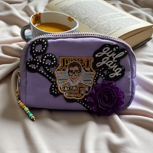 Può includere: Un beauty case lavanda con le parole "girl gang" e "Dissent". La borsa ha una chiusura con cerniera e un tirante con perline. Una rosa di stoffa viola e abbellimenti in pizzo nero decorano la borsa. Una tazza di tè e un libro sono sullo sfondo.