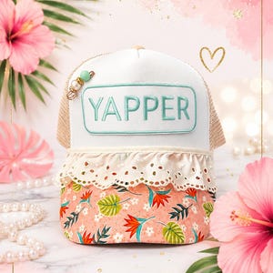 May include: A white trucker hat featuring a rectangular patch with the word YAPPER in teal embroidery. The hat includes a cream-colored eyelet lace trim, a peach-colored brim with a tropical floral pattern, and a small decorative bead charm.