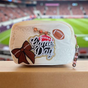 以下が含まれることがあります： クリーム色のジッパー付きポーチ、アメフトテーマ。茶色のリボン、フットボールのアップリケ、金と白で「Game Day」の文字が特徴。装飾ビーズとレースのディテールで飾られています。小物を持ち運ぶのに最適です。
