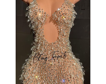 May include: A sheer, nude mini dress completely covered in rhinestones. The dress has a plunging neckline and a fitted silhouette. The dress is labeled "Bling Imports" on the bottom.