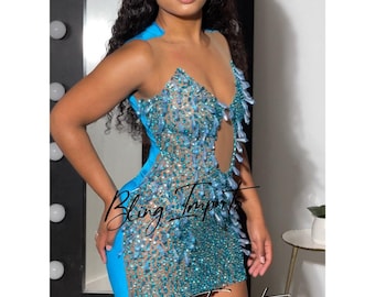 Puede incluir: Un vestido corto turquesa con un corpiño transparente cubierto de pedrería azul y transparente. El vestido tiene un escote pronunciado y una silueta ajustada.
