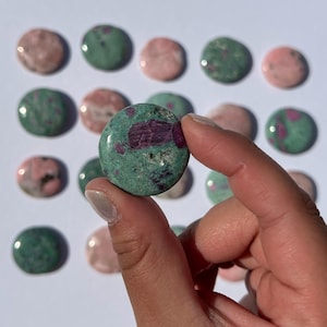 May include: A collection of polished gemstones in shades of pink and green, with one held in a hand. The held stone is a vibrant green with deep red inclusions. Other stones are scattered in the background, showcasing variations in color and pattern.