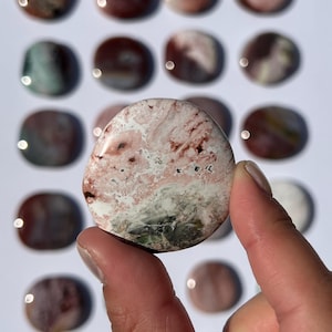 May include: A polished, oval-shaped stone with a mix of colors, including pink, white, and brown. The stone is held in a person's hand, with several other similar stones in the background. The stones appear to be natural gemstones.