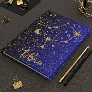 Pode incluir: Um caderno azul com fundo de glitter dourado e um design de constelação dourada. A constelação é etiquetada como "Libra".