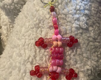 May include: A pink and red beaded gecko keychain. The gecko is made of small, round beads and has a yellow string attached to a silver key ring.