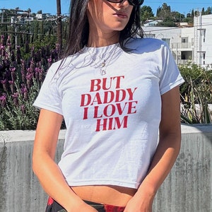 Op de afbeelding: Wit kort T-shirt met rode tekst die "BUT DADDY I LOVE HIM" zegt. De persoon draagt een bloemenrok.
