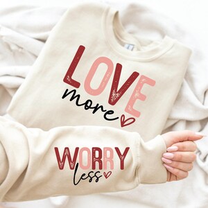 Peut inclure: Un sweat-shirt beige avec le texte "LOVE more" en lettres roses et rouges avec un petit cœur sur le devant et le texte "WORRY less" en lettres roses et rouges avec un petit cœur sur la manche.
