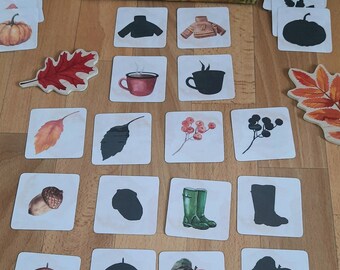 May include: A set of 16 matching cards featuring various autumn-themed images and their corresponding silhouettes. The cards are arranged in a grid on a wooden surface. The images include a red leaf, a brown acorn, a green boot, a black umbrella, a red apple, and a brown cup of coffee.