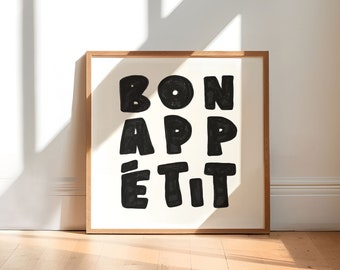 以下が含まれることがあります： クリーム色の背景に、太字の黒い文字で「BON APPÉTIT」と書かれた額入りのアートプリント。木製のフレームは、白い壁に立てかけられた木製の床に置かれています。日光が壁と床に影を落としています。
