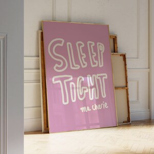 May include: A pink framed poster with white text that reads "SLEEP TIGHT ma chérie" hanging on a white wall in a room with white trim.