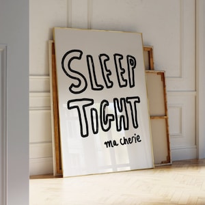 May include: A white framed poster with black text that reads "SLEEP TIGHT ma cherie". The poster is leaning against a white wall and a wooden floor.