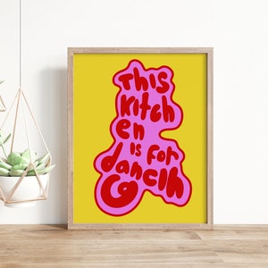 Puede incluir: Un fondo amarillo con un gráfico rosa y rojo que dice "This kitchen is for dancing". El gráfico está en un estilo moderno, dibujado a mano. El gráfico está enmarcado en un marco de madera claro.