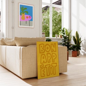 May include: A yellow poster with the text "BON APP&Eacute;TIT" in a bold, orange font. The poster is leaning against a beige couch in a living room.