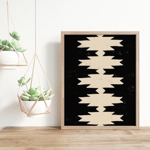May include: A framed print with a black background and a beige geometric pattern. The print is hanging on a white wall above a wooden table with two hanging planters with succulents.