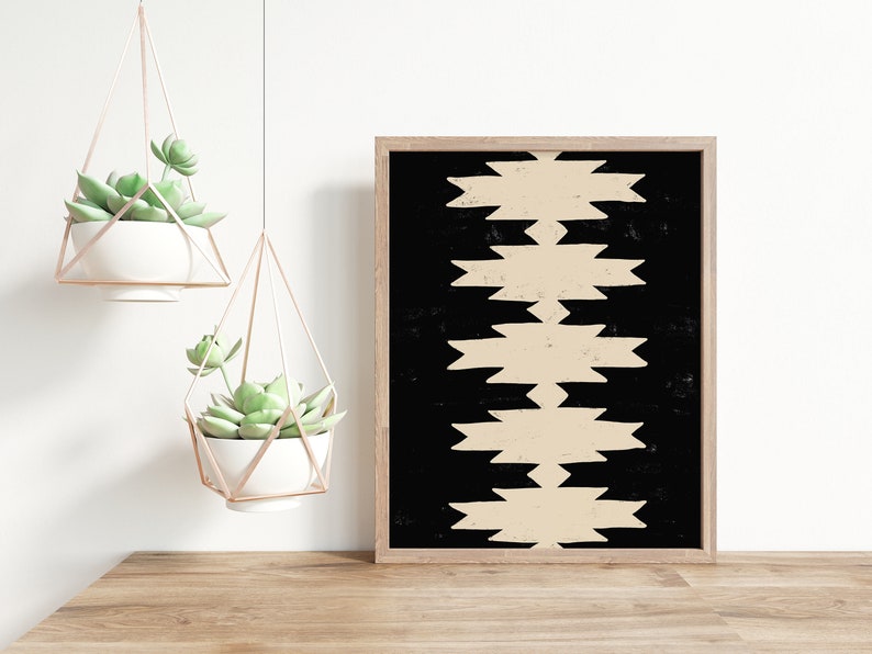 May include: A framed print with a black background and a beige geometric pattern. The print is hanging on a white wall above a wooden table with two hanging planters with succulents.