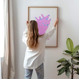 May include: A person hangs a framed print on a white wall. The print features a pink and blue line drawing of two hands clasped together.