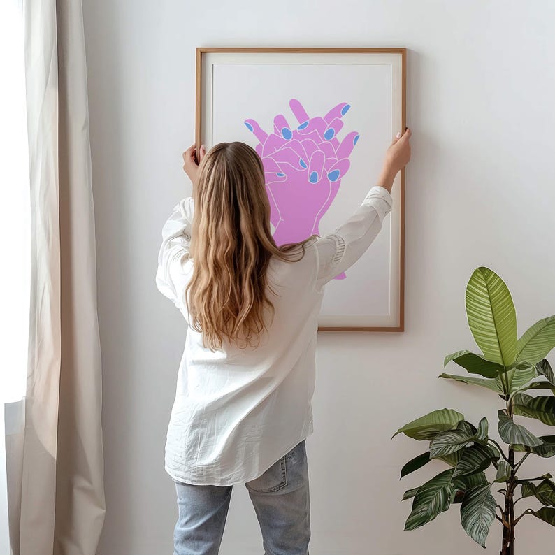 May include: A person hangs a framed print on a white wall. The print features a pink and blue line drawing of two hands clasped together.