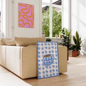 May include: A blue and white floral print poster featuring a blue teacup and saucer with brown tea inside. The teacup has a white floral pattern. The poster is leaning against a beige sofa.