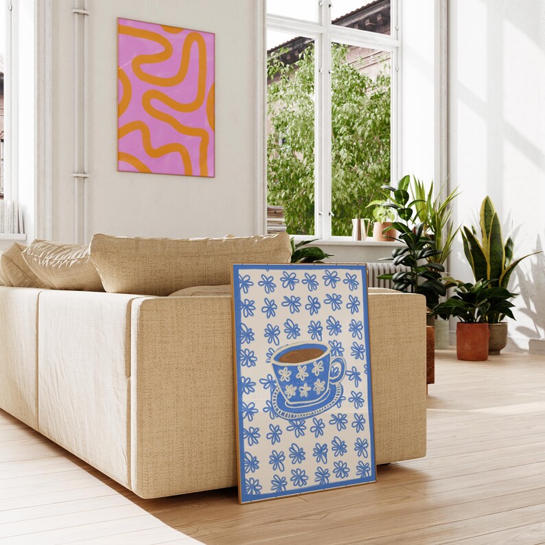 May include: A blue and white floral print poster featuring a blue teacup and saucer with brown tea inside. The teacup has a white floral pattern. The poster is leaning against a beige sofa.