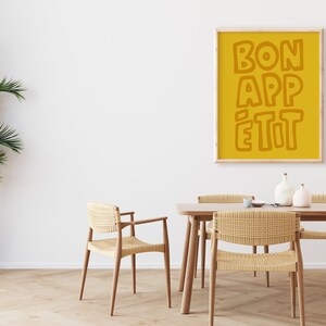 May include: A yellow poster with the text "BON APP&Eacute;TIT" in a bold, orange font hangs on a white wall above a wooden dining table with four woven chairs.