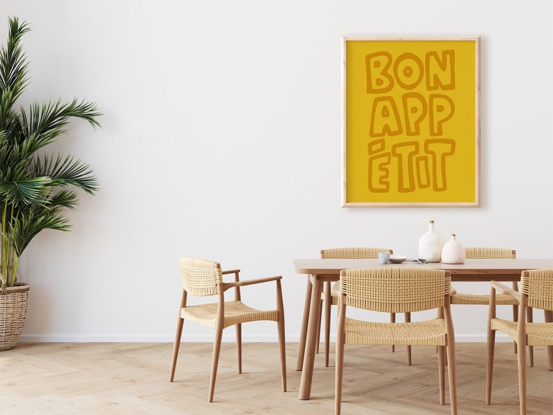 May include: A yellow poster with the text "BON APP&Eacute;TIT" in a bold, orange font hangs on a white wall above a wooden dining table with four woven chairs.