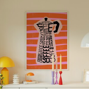 May include: A framed print of a pink coffee pot with the word "coffee" repeated in black text on an orange and pink striped background. Three candlesticks with candles are on a white surface in front of the print.