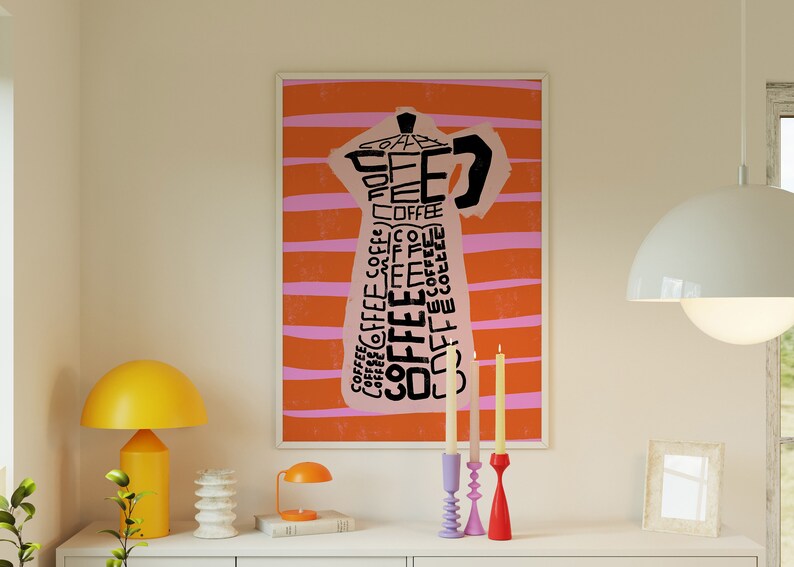 May include: A framed print of a pink coffee pot with the word "coffee" repeated in black text on an orange and pink striped background. Three candlesticks with candles are on a white surface in front of the print.