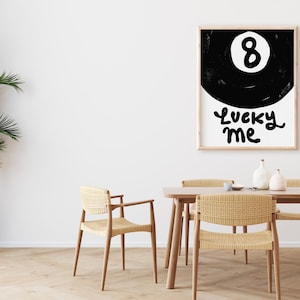 May include: Black and white illustration of a pool ball with the number 8 on it. The text "Lucky Me" is written below the ball. The illustration is framed and hanging on a white wall above a wooden table with chairs.