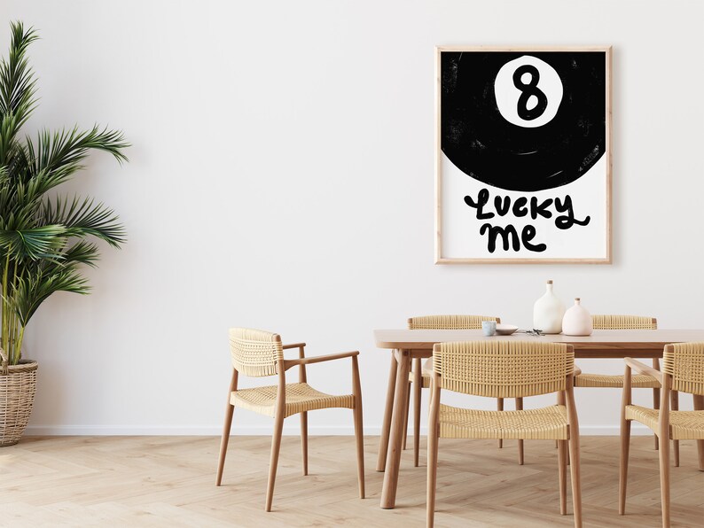 May include: Black and white illustration of a pool ball with the number 8 on it. The text "Lucky Me" is written below the ball. The illustration is framed and hanging on a white wall above a wooden table with chairs.