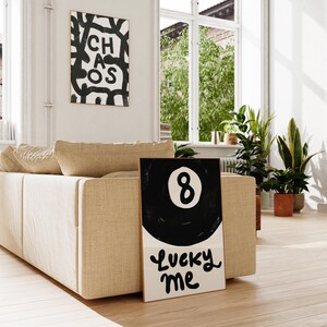 May include: A black and white illustration of a pool ball with the number 8 on it. The text "Lucky Me" is written below the ball. The illustration is framed and leaning against a beige couch.