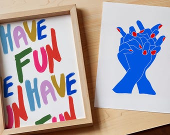 May include: Two framed prints with colourful designs. The first print has the words "HAVE FUN WE HAVE FUN" written in a variety of colours. The second print features two blue hands with red fingernails holding each other.