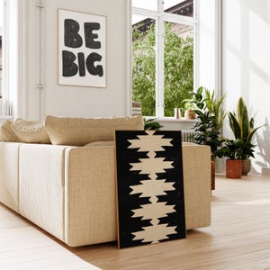May include: A beige sofa with a black and white geometric print artwork leaning against it. The artwork is in a wooden frame. The sofa is in a room with a window and plants.