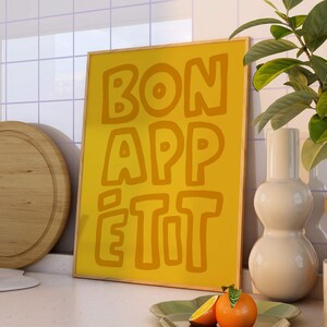 May include: A yellow framed poster with the text "BON APP&Eacute;TIT" in a bold, outlined font. The poster is hanging on a white tiled wall in a kitchen setting.