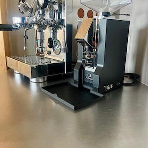 Può includere: Una macchina per espresso cromata con portafiltro e un macinacaffè nero con tramoggia trasparente e vassoio con etichetta Silenzio. Il macinacaffè presenta un pannello frontale metallico, una manopola di regolazione e un interruttore laterale.