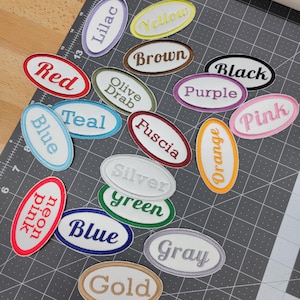 May include: A collection of oval-shaped embroidered patches in various colors, including red, blue, and yellow. Each patch features a different color name in a matching font. The patches are arranged on a gray cutting mat.