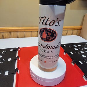 May include: White stainless steel travel mug with a black lid. The mug has a Tito's Handmade Vodka logo with the text "Award Winning" and "Handmade Vodka".