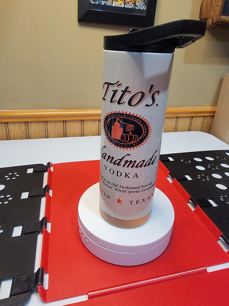 May include: White stainless steel travel mug with a black lid. The mug has a Tito's Handmade Vodka logo with the text "Award Winning" and "Handmade Vodka".
