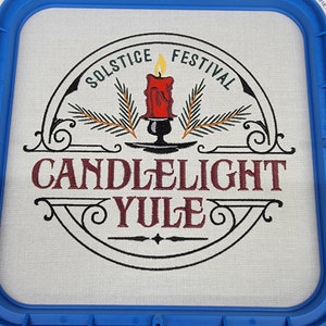 May include: An embroidered design on white fabric in a blue embroidery hoop. The design features a candle with flames, pine branches, and the text "Solstice Festival Candlelight Yule".