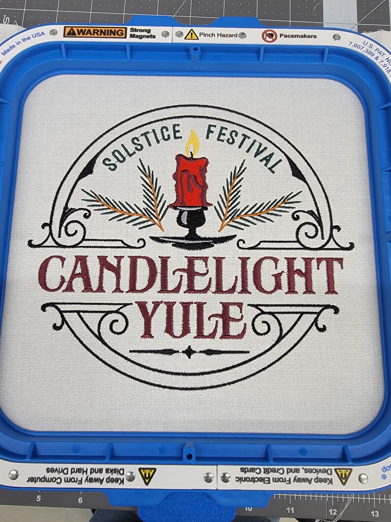 May include: An embroidered design on white fabric in a blue embroidery hoop. The design features a candle with flames, pine branches, and the text "Solstice Festival Candlelight Yule".