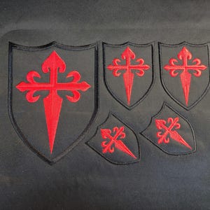 May include: Five black fabric patches shaped like shields, each featuring a vibrant red embroidered cross with pointed ends. The largest patch is on the left, with two medium-sized patches above and two smaller patches below, all on a black background.