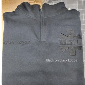 May include: Black quarter-zip sweatshirt with "Paramedic" emblem and "Jaylen Hoyer" text. Features black-on-black logos and a zipper closure. The sweatshirt is folded on a gray surface.