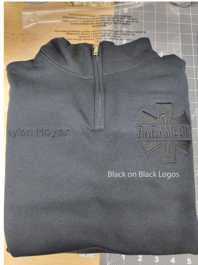May include: Black quarter-zip sweatshirt with "Paramedic" emblem and "Jaylen Hoyer" text. Features black-on-black logos and a zipper closure. The sweatshirt is folded on a gray surface.